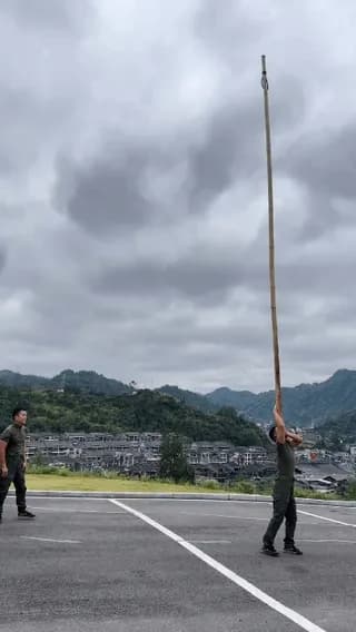 Holding a bamboo pole on his shoulder while another performs acrobatics at the top.