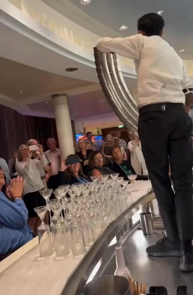 A bartender pours cocktails through a chain of connected metal cups, filling multiple glasses at once.