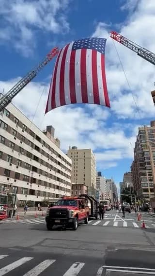 NYC firefighters honoring Father Mychal Judge, the first certified fatality of 9/11, by suspending a massive flag over the remembrance walk.